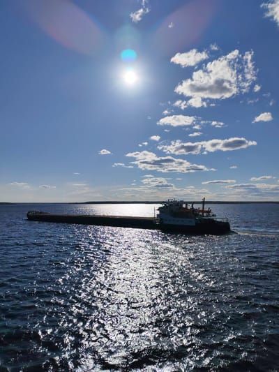 A barge is moving on a sunlit body of water beneath a clear blue sky dotted with white clouds. The barge is dark in color and appears to be carrying a load. The water is choppy, with sunlight reflecting off the surface creating bright sparkles. The horizon shows land in the distance. The photograph was taken in Rechnaya Sosnovka, Russia.