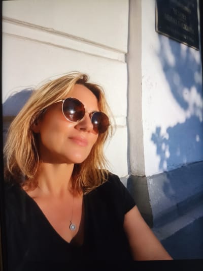 In a medium shot, a woman with blonde hair and sunglasses is positioned in the foreground, facing towards the left side of the frame. She is wearing a black v-neck top and a pendant necklace. The background consists of a textured white wall with horizontal lines and a darker rectangular plaque with text that appears to be in Cyrillic script. To the right of the woman, the wall transitions into a gray, stepped surface. Sunlight casts dappled shadows on the wall, suggesting an outdoor setting during the day. The overall scene is brightly lit, with a strong contrast between light and shadow. The image is likely taken in Chekhov-2, Russia, as indicated by the plaque which contains text referencing A.P. Chekhov.