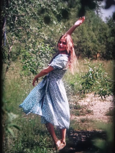 The media file shows a young woman in a light blue and white floral dress, barefoot, posing outdoors in a natural setting. She is in motion, with her left arm raised high and her body angled as if dancing or twirling. Her hair is long and a reddish-brown color, falling around her shoulders. The background consists of lush green foliage, trees, and dry grass, suggesting a warm, possibly summer day. Sunlight filters through the leaves, creating dappled light on the ground. The scene evokes a feeling of freedom and joy. The location context is Serov, Russia. There is no visible text in the media file.