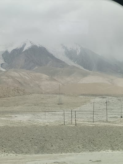 A vast, desolate landscape under a cloudy sky dominates the scene. In the distance, snow-capped mountains rise, their peaks shrouded in mist. The terrain in the foreground is arid and barren, with rolling, sandy hills and a rocky, gravelly plain. Several utility poles with wires are scattered across the landscape, suggesting a remote, possibly undeveloped area. The overall impression is one of stark, natural beauty and isolation. This image depicts the outskirts of Bulungkol Township, China, a region characterized by its harsh, mountainous terrain and sparse vegetation.
