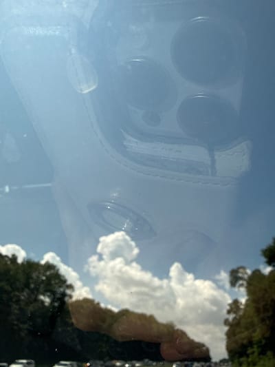 This is a close-up, angled, indoor shot of a car's windshield. The windshield reflects the exterior, showing a bright, sunny sky with fluffy white clouds. Trees and what appear to be other cars are visible in the distance, suggesting a road or parking lot environment. The reflection also shows a phone with multiple cameras, likely the device used to take the photo. The overall impression is a sunny day viewed through a car window. The location is the Village of Lake Success, United States of America.