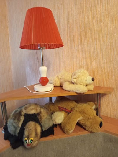 The image shows a collection of stuffed animals on a wooden shelf against a textured, light-brown wall. On the left, a light-colored teddy bear is propped up against the shelf. To its right, a larger brown teddy bear with a red and black striped ribbon around its neck is also resting on the shelf. Below the brown teddy bear, a black and brown stuffed animal, resembling a cat with large, round eyes, is positioned. A red lampshade with a metal stem and a white and red knob is attached to the wall on the left side of the image. The scene appears to be indoors, possibly in a child's room or a decorative display. The lighting suggests it could be daytime, with no indication of specific weather conditions. There is no visible text in the image. The overall impression is one of a cozy and playful arrangement of toys.
