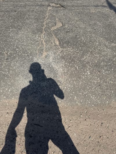 In Serebryany Bor, Russia, a shadow of a person is cast on a sunlit asphalt surface. The shadow, elongated and dark, covers a significant portion of the frame, obscuring the details of the ground beneath. The asphalt itself appears aged and worn, with visible cracks and patches of lighter material. Small pebbles and debris are scattered across the surface, particularly in the lower right and left portions of the image. The lighting suggests it is daytime, with the sun positioned to create the prominent shadow.