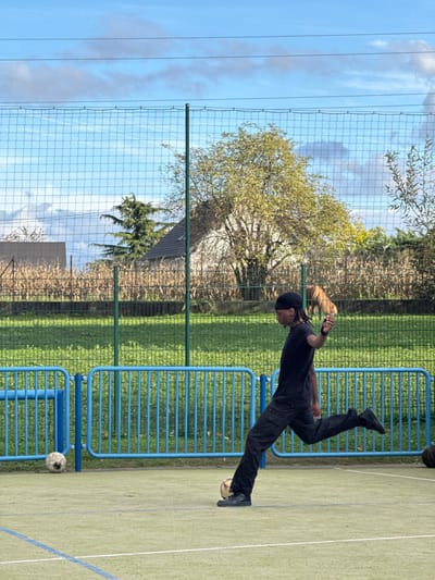 The image depicts an outdoor sports court in Niederhausbergen, France, where a male individual is captured in motion, kicking a light-colored football with his right foot. He has dark skin, wears a black durag, a black t-shirt, black trousers, and black sneakers; his dreadlocks are visible. Another dirty white football rests on the court to the left. The playing surface is green with visible white and blue court lines. A blue metal barrier runs across the midground, separating the court from a grassy field. Beyond the field and a green mesh fence, the background features a field of dry, light brown crops, a deciduous tree with light green/yellow foliage, a dark green coniferous tree, and part of a building with a dark-tiled roof. The sky is blue with scattered white clouds and faint overhead power lines.