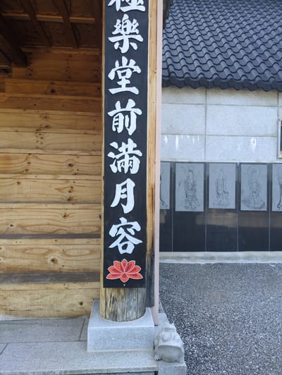 This forensic description details an outdoor scene within a Buddhist temple complex, specified as being located in Godang-ri, South Korea. The image is captured at eye-level, under bright daylight, revealing architectural elements and signage.

In the left foreground, a substantial vertical wooden post stands, exhibiting a weathered texture and visible wood grain. Attached to the post is a prominent black rectangular sign, oriented vertically, featuring large white Japanese characters. The characters read from top to bottom as "極楽堂前満月容" (Gokurakudō-mae Mangetsu-yō), which translates to "In front of Pure Land Hall, Full Moon Countenance/Appearance." Below the inscription, a stylized red lotus flower with lighter red and orange accents is depicted. The base of the wooden post is set into a light grey, polished granite foundation.

Immediately to the left of the signpost, a wooden building facade is visible. It is constructed from horizontal planks of light-colored wood, displaying natural grain patterns and various knots. The underside of a dark, exposed roof structure is discernible at the top left edge of the frame, above the wooden facade.

To the right of the signpost, another building is present, characterized by a dark, corrugated or wave-patterned tiled roof that extends across the upper right portion of the image. Below the roofline, a wall section is composed of light grey, square or rectangular stone panels, showing slight discoloration or marks. Further down, directly below this lighter wall, a series of darker, vertical rectangular panels are affixed. These black panels feature engraved or etched images of standing Buddhist figures, each with a halo and detailed robes. Four such figures are clearly visible across the frame. The wall concludes with a dark, low-lying stone base at ground level.

The ground surface in the middle and right foreground consists of a speckled, dark grey aggregate paving, suggesting gravel or textured concrete. Adjacent to the signpost's base, a smoother, lighter grey stone slab forms a walkway. A small, light grey stone statue of a frog is situated on the edge of this stone slab in the bottom right corner, facing towards the left. There are no people present in the scene.