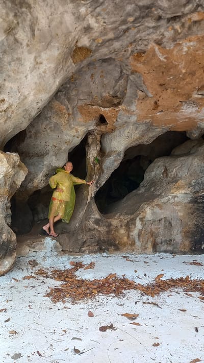 A medium shot depicts a woman in a yellow raincoat and sandals inside a cavern. She's reaching upwards towards a rocky overhang. The cavern's walls are composed of textured rock formations with hues of gray, beige, and brown. The floor outside the cavern entrance is white with scattered brown leaves. The location is Thap Put, Thailand.