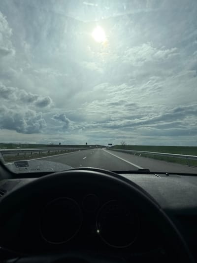 The image shows a view from inside a vehicle, likely a car, driving on a highway. The steering wheel is visible on the right side of the frame, suggesting a left-hand drive vehicle. The road ahead is straight, with a white line marking the lanes. A metal guardrail separates the highway from a grassy area on the right. The sky is overcast with clouds, and the sun is shining brightly in the upper left corner of the frame. The location is unspecified. No people, subjects, actions or interactions are visible. There are no references to Malenovo, Bulgaria.