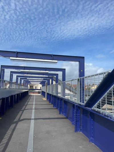 The image depicts an elevated pedestrian walkway or bridge, viewed from a low angle, looking forward along its length. When oriented correctly, the scene is bathed in bright daylight under a clear blue sky sparsely dotted with small, rippled clouds.

The most prominent features are a series of royal blue metallic structural beams that form a repeating archway or frame over the walkway. These robust beams are bolted together with visible rivets and extend into the distance, creating a sense of perspective. On the right side of the walkway, a silver metallic mesh railing runs parallel to the path, affixed to the blue structure. On the left side, several long, white fluorescent light fixtures are suspended from the blue overhead beams.

The walkway surface is dark grey asphalt, featuring a distinct light grey tactile paving strip running down its center. Strong, elongated shadows from the overhead structure and the right-side railing are cast across the path, indicating bright sunlight. In the background, through the framework of the structure and above the railing, the rooftops and upper parts of various buildings are visible, suggesting an urban or semi-urban setting.

No people are visible, and there is no specific activity taking place. A small yellow square with the number "22" in black is affixed to one of the blue structural beams. The modern, industrial design of the structure points to contemporary infrastructure, likely a public thoroughfare such as a footbridge or platform access.