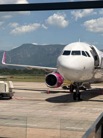 An airplane with pink accents is parked on an airport tarmac. The plane's nose and cockpit windows are visible, as is its left engine nacelle, which is also accented in pink. The wingtip of the plane shows the airline's branding. The tarmac is marked with yellow and white lines, and a red hose runs from a service vehicle to the plane. In the background, a range of mountains forms the horizon under a partly cloudy blue sky. The scene appears to be captured from an elevated vantage point, possibly a terminal window, as a dark horizontal beam is present at the top of the frame. This scene is located at Tirana International Airport Nënë Tereza (TIA) in Kruja, Albania.