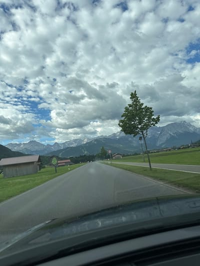 The image shows a scenic view from a car driving on a road in Farchant, Germany. The road leads towards a majestic mountain range under a dramatic sky filled with clouds. On the left side of the road, there are a couple of rustic wooden sheds with red roofs. A sign with green text is visible near one of the sheds. A single tree stands tall on the right side of the road, with a speed limit sign (70) nearby. The landscape is characterized by green fields and distant villages nestled at the foothills of the snow-capped mountains. The perspective suggests the photo was taken from the driver's seat, capturing the road ahead and the surrounding natural beauty.