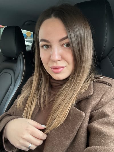 A woman is pictured in the interior of a car. She has light brown hair and is wearing a brown turtleneck sweater and a brown wool coat. She is wearing a silver ring on her ring finger. A black leather car seat is visible. The location cannot be determined from the image.