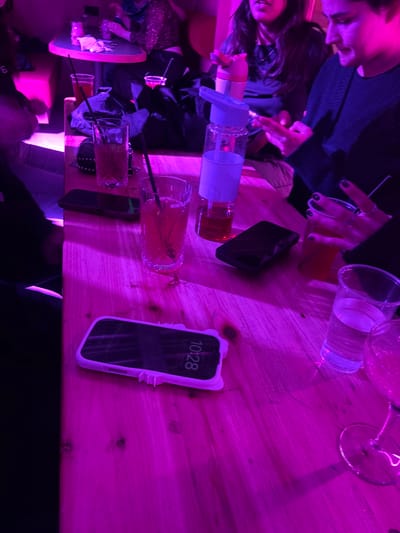 A table is illuminated by purple light, cluttered with drinks in glasses and plastic cups, a hand sanitizer dispenser, and two smartphones. One phone displays the time as 10:28 and is in a decorative white case. Two people are partially visible, one on either side of the table. Black clothing and accessories are scattered around. The table appears to be made of light-colored wood.