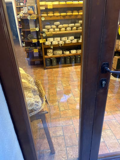 This image captures the interior of a traditional Italian food shop, likely a deli or "formaggeria," viewed through a glass door or window. The perspective is angled, looking down into the shop's brightly lit display area.

The shop's left and central sections are dominated by multi-tiered wooden shelves laden with a wide assortment of cheeses. Numerous cheese wheels and smaller rounds, varying in size and maturity, are neatly arranged. Many cheeses feature yellow labels indicating varieties such as "Pecorino," "Pecorino Fresco," "Pecorino Semistagionato," "Pecorino Stagionato," "Caciotta," and "Ricotta Salata." Some tags also include "Pienza," reinforcing the provided location context. Small bunches of dark grapes are nestled among some of the cheeses as decoration. Further into the shop, on the right, shelves display stacks of black rectangular packages, possibly other specialty food items.

In the foreground, to the right, a large wicker basket is prominently featured, overflowing with copious amounts of light-colored, thin-strand fresh pasta, reminiscent of tagliolini or spaghetti. The shop floor, visible through the display, is covered with warm, earthy-toned rectangular tiles laid in a running bond pattern.

The top and right edges of the image are framed by a dark wooden door or window frame with a glass panel, which causes subtle reflections. A keyhole mechanism is visible on the wooden frame. The scene is static, showing no people or specific activity, focusing solely on the artisanal food products. The array of cheeses, particularly Pecorino, and fresh pasta, combined with the traditional shop aesthetic, perfectly aligns with a specialty food store in Pienza, Italy, a town celebrated for its local cheese production.