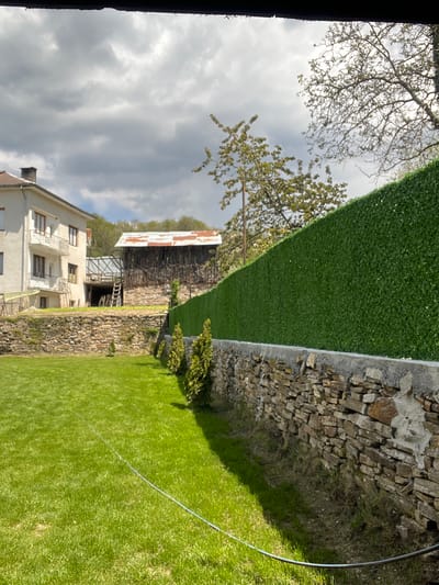 This image captures a quiet, sunlit rural or suburban landscape in Kribul, Bulgaria, viewed under an overcast yet bright sky. The scene is static, with no visible human activity.

In the foreground, a lush, well-maintained green lawn stretches across the lower and right portions of the frame. A thin, light-colored cable or hose lies on the grass, extending from the right edge towards the center. Separating the lawn from a lower area is a sturdy stone wall, built with natural, irregularly shaped rocks, running horizontally across the mid-ground. Perched atop this stone wall is a neat, uniform artificial green hedge or fence, providing a verdant backdrop.

To the left of the stone wall and below the level of the lawn, a multi-story house with light-colored facades and multiple balconies is visible. Further back and to the right of this house, behind the artificial hedge, stands a rustic, older building, possibly a barn or outbuilding. This structure features exposed wooden beams and a large, partially rusted corrugated metal roof. A few small, leafy bushes or trees are clustered near the stone wall on the lower ground level.

The background reveals a mix of trees; some are full of green leaves, while others show more bare branches against the sky. The sky above is predominantly cloudy, with varying shades of gray and white, suggesting an overcast day with some bright light filtering through. A dark, vertical frame element partially obscures the far left edge of the image. No text is visible anywhere in the scene.