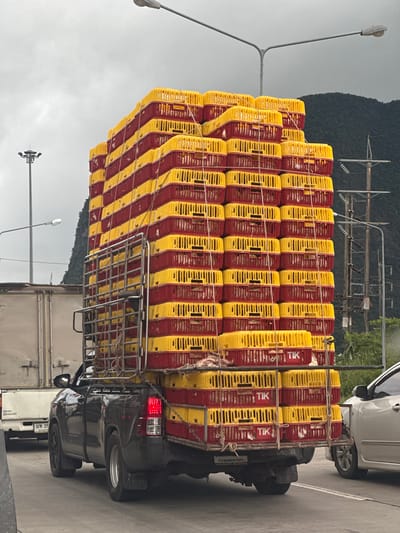 A black pickup truck is seen on a road in Phang-nga, Thailand, overloaded with stacked yellow and red chicken crates. The crates are arranged in a towering formation, filling the truck bed and extending high above it. The truck is in motion, indicated by the blurred surroundings and the position of the wheels. A light-colored truck is to the left of the pickup, and a light-colored sedan is to the right, suggesting a traffic situation. The background features power lines, a hill, and an overcast sky, typical of the tropical climate in Phang-nga. The crates appear to be for transporting live poultry, with visible openings and some birds peeking out. The scene captures a moment of everyday logistics and transportation in a Thai city.