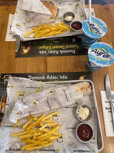 Two meals are displayed on a wooden table, each served on a metal tray lined with branded paper. Each tray holds a doner kebab or wrap, a generous serving of french fries, and two small metal cups containing ketchup and mayonnaise. Utensils, a knife on one tray and a fork on the other, are also present.
Accompanying each meal is a plastic cup of Ayran, a traditional Turkish yogurt drink, identifiable by the blue and white "sütaş Ayran" branding. The branded paper liners within the trays prominently feature the text "Bursa Kebap Evi" and the initials "BKE," indicating the restaurant's name.
Two paper table runners or placemats are visible on the table. The one on the left displays the phrase "Dominik Adası'nda Lezzet Devam Ediyor..." (which translates to "Taste Continues on Dominik Island..."), while the one on the right shows "Dominik Adası'nda". The overall setup suggests a casual dining experience, likely at a fast-food establishment or a restaurant specializing in Turkish cuisine. The drinks and restaurant name strongly imply a Turkish setting, consistent with the given location context of Bahçelievler, Turkey. No people are visible, and the time of day and weather cannot be determined.