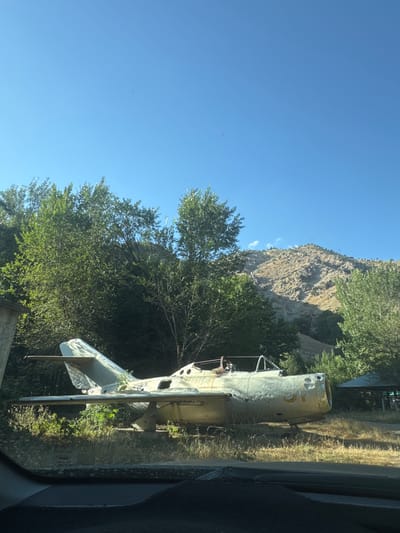 An old, weathered, light-colored jet airplane is parked on grassy terrain in Mirta, Tajikistan. The plane, which appears to be a Soviet-era fighter jet, is missing its canopy and shows signs of wear and tear, including visible dents and paint chips. It is situated amidst dense green foliage and trees, with a rugged, rocky mountainside in the background under a clear blue sky. The foreground shows the interior of a vehicle, suggesting the photo was taken from a car.