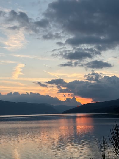 The image captures a panoramic view of the Dospat Dam in Dospat, Bulgaria, at sunset. The sky, occupying the upper portion of the frame, transitions from pale blue and light yellow in the upper left to darker grey and blue clouds in the upper right. Several white contrails are visible, with a prominent one extending diagonally from left-center to right-center, and another fainter one in the upper left. The setting sun, obscured by clouds and the distant horizon, casts intense orange and red light low on the horizon, particularly on the right side, illuminating the undersides of dark, volumetric clouds. A continuous silhouette of undulating mountains or hills forms the horizon line, separating the sky from the expansive body of water. The calm surface of the Dospat Dam reflects the vibrant orange and red hues of the sunset, creating shimmering patterns across the water, which also exhibits subtle, parallel ripples. In the mid-distance on the water, a small, dark cluster of objects, possibly birds, is visible. The immediate foreground on the lower right features a dark, silhouetted cluster of vegetation, likely tall grasses or reeds, extending into the frame. No human figures or discernible man-made structures other than the contrails are present.