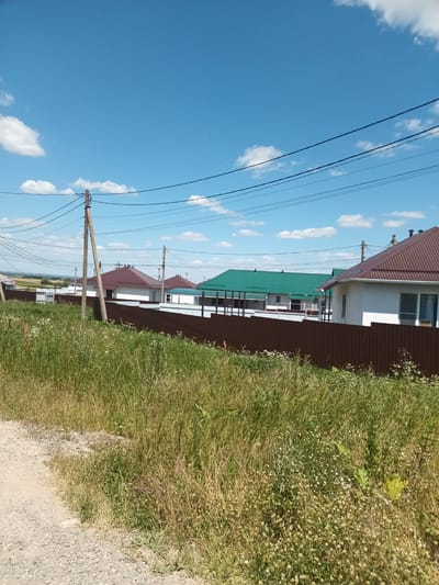 This image depicts a residential area in Krymsk, Russia, under a bright blue sky with scattered white clouds. In the foreground, a dirt road or path borders a field of tall, dry grass with some wildflowers. Several houses with brown metal roofs are visible behind a brown corrugated metal fence. One house on the right is white with a brown trim around the roofline. Another house to the left has a green metal roof. Power lines are strung across the sky, supported by wooden utility poles. The scene appears to be a quiet, rural or suburban setting.