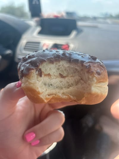 A close-up shot shows a person's hand holding a partially eaten pastry. The pastry appears to be a type of fried dough, coated in dark chocolate with small chocolate chips or pieces embedded in the coating. The interior of the pastry is visible, showing a light, airy texture. The hand holding the pastry has bright pink nail polish on its fingernails.
In the background, the scene is blurred, but suggests an outdoor setting. There are hints of a light-colored vehicle or structure, possibly with some red accents, and a bright, possibly sunny sky. The overall lighting suggests it is daytime. The location context of Tsalapitsa, Bulgaria, is noted but not directly visible in the image. No visible text is present. The activity appears to be someone enjoying a snack.