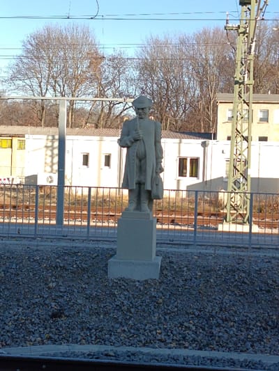 The image displays a daylight scene at the Altenburg train station in Altenburg, Germany. In the mid-ground, a light-colored stone statue of a standing male figure is centrally positioned. The figure wears a brimmed hat, a long coat, breeches, and stockings, holding what appears to be a walking stick or pipe in its right hand and a bag or satchel in its left hand. The statue rests on a two-tiered rectangular pedestal of similar light-colored stone.
The immediate foreground consists of grey gravel. Beyond the statue and gravel, a grey metal barrier fence with vertical bars and horizontal rails separates this area from multiple parallel railroad tracks set on a ballast bed. Overhead electrical catenary lines stretch across the top portion of the frame. To the right of the statue, a tall, green-painted metal lattice mast stands beside the tracks.
In the background, behind the tracks and fence, several buildings are visible. A prominent, long, single-story building with a light-colored facade and a brown tiled roof features multiple rectangular windows, some reflecting light. An external air conditioning unit is mounted on its side. To the far left, a portion of another building with distinctive yellow-tinted windows is visible. To the far right, a two-story building with a brown roof is partially obscured by the lattice mast. A dense cluster of bare deciduous trees forms a natural backdrop behind the buildings. The sky is clear and blue, and the scene is brightly illuminated by sunlight originating from the right, casting discernible shadows.