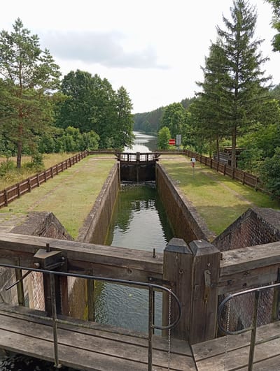 The image shows a canal lock in Płaska, Poland. The lock has brick walls and wooden gates. The water level in the lock is lower than the surrounding canal, suggesting that the gates have just been opened or are about to be closed. There is a wooden walkway on either side of the lock, and a wooden fence runs along the embankment. Trees and vegetation surround the lock, and a body of water can be seen in the distance. The sky is cloudy. There are no people or animals visible in the image. The scene appears to be a quiet and natural setting.