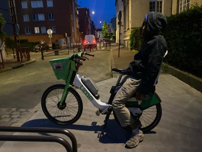 The image shows a person seated on a Lime electric bicycle on a street in Le Pré-Saint-Gervais, France. The individual is wearing a dark hoodie and light-colored pants, with a pair of sneakers visible at the bottom of the frame. The Lime bike is parked on the sidewalk near a metal railing. A multistory brick building is located on the left side of the frame, with windows visible on each story. A white van with its brake lights activated is visible in the background. Streetlights are present on the road, illuminating the scene. A lighter brick building is visible on the right side of the frame, partially obscured by dense foliage. The sky is dark blue, suggesting that it's dusk or night.