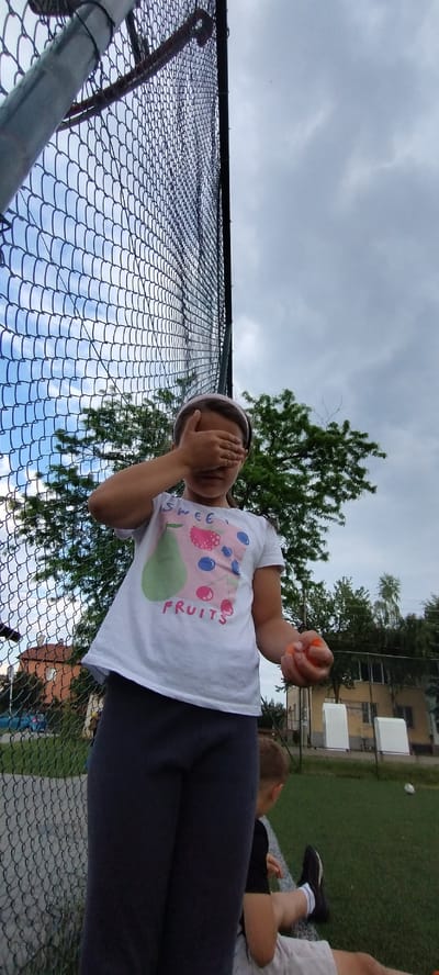 A low-angle, wide-shot photograph captures an outdoor scene at a sports or recreational area in Gara Elin Pelin, Bulgaria, under an overcast sky. A chain-link fence dominates the left and upper portions of the frame, serving as a prominent visual element and partially obscuring the background.
In the foreground, slightly left of center, a young girl stands facing the camera. She wears a white t-shirt with a "SWEET FRUITS" graphic depicting a green pear, red berries, and blue berries. Dark grey leggings cover her lower body. A light pink headband is visible on her head. Her right hand is held horizontally across her face, completely covering her eyes. In her left hand, she holds a small, orange, spherical object.
To the right of the girl and in the lower right quadrant, a young boy is seated on artificial green turf. He is viewed from behind and slightly to the side, wearing a dark t-shirt, light-colored shorts, black sneakers, and white socks. A white line is visible on the artificial turf near where he is seated.
Beyond the chain-link fence, the background reveals a mix of natural and man-made elements. Green foliage from trees is visible, with some patches of lighter blue sky breaking through the clouds. In the mid-ground, various buildings are discernible, including a light-colored structure with white accents. A blue vehicle is partially visible through the fence on the left side of the background. Two white, rectangular structures, possibly goal posts or field markers, are positioned on the artificial turf in the distant background, to the right of the buildings. A small white ball rests on the artificial turf further back. The fence structure includes a grey metallic post on the far left and a reddish-brown hooked bar extending downwards from the top of the fence line.