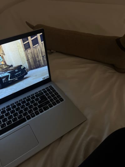 The image depicts a laptop placed on a bed with white sheets. The laptop screen displays a racing game, featuring a black Lexus car parked in front of a wooden garage door. The keyboard is visible, with keys displaying both Latin and Cyrillic characters. A long, textured brown pillow is positioned behind the laptop. The scene is dimly lit, suggesting it is nighttime or the room is dark. The location in the city of Balbriggan, Ireland, is not discernible from the image.