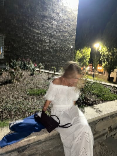 A nighttime photograph taken in Mogadouro, Portugal, captures a single individual seated on a stone ledge. The person, with blonde hair blurred by motion, wears a white off-the-shoulder eyelet-embroidered top and a matching long skirt. A thin pink band is visible on their right wrist. A dark brown, possibly woven, handbag with a strap rests on the ledge next to the person. To the left of the individual, a blue garment, appearing to be a hoodie with partially visible white text, is placed on the stone surface. The background features a tall, dark building wall constructed from textured, rough-hewn stone on the left. To the right, beyond a low planter containing sparse vegetation, a bright streetlamp casts an orange-yellow glow on green-foliaged trees and distant buildings, all under a dark night sky.