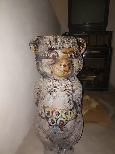 A ceramic sculpture of a bear-like creature, standing approximately two feet tall, is the central focus of this indoor scene. The sculpture has a textured, glazed finish with shades of brown, white, and blue, and features large, round eyes. A band of multiple circular patterns, resembling eyes or targets with red accents, encircles the creature's waist. The sculpture is positioned in front of a white wall on the left and a dark entertainment center on the right. The entertainment center consists of a black television set on top, with a microwave oven below it. The room appears to be dimly lit, with the primary light source possibly coming from the left side of the frame, casting a soft shadow on the wall. A light-colored, amorphous object, possibly a piece of fabric or paper, is visible on a wooden surface behind the sculpture. The location is identified as Aygezard, Armenia.