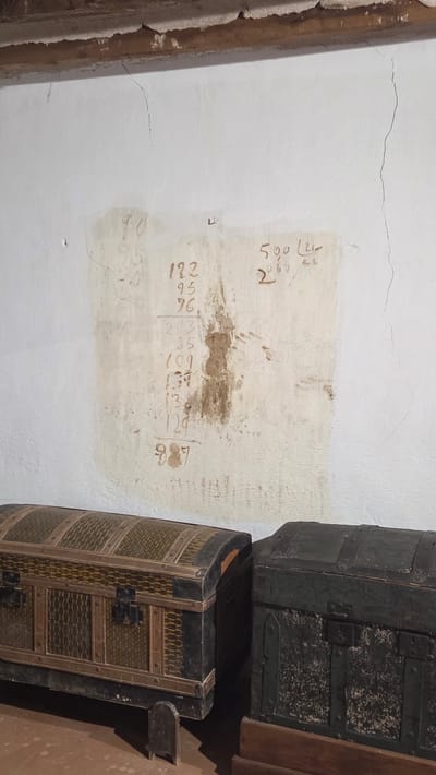 The image shows a section of an interior wall in Altura, Spain. The wall is painted white and has a rectangular patch of a different shade of white. The patch contains faded numbers and calculations written in brown ink or paint, suggesting a historical or archival context. A dark-colored wooden beam is visible on the left edge of the image. Two antique chests or trunks are stacked against the right side of the wall. The top trunk is dark green with metal hardware, while the bottom trunk is a curved, barrel-shaped piece with decorative details and metal fixtures. The environment suggests a historical building or museum setting.