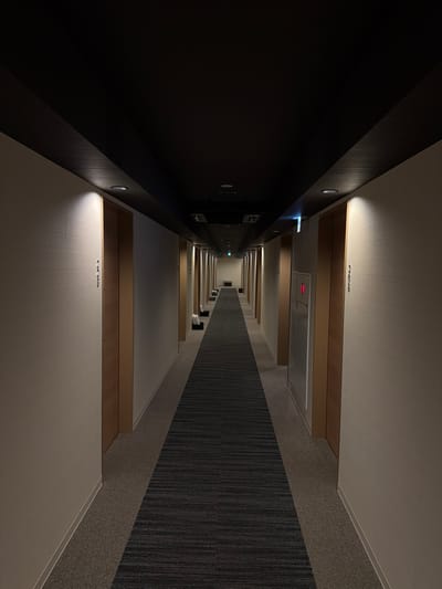 This image displays a long, empty interior hallway within a building in Kutchan, Japan. The perspective is centered, looking down the corridor towards a distant end wall featuring a dark, low bench. The ceiling is uniformly dark brown to black, housing multiple circular recessed spotlights that cast pools of warm light onto the light beige, textured walls. A dark central beam extends along the length of the ceiling. Numerous light-colored wooden doors, each with vertical trim, line both sides of the hallway at regular intervals. Japanese characters and numerical sequences, such as "A-F A-F B-C-A" and "42555," are vertically inscribed on the wall surfaces adjacent to some doors. On the right side, a silver-colored fire extinguisher cabinet with a red accent is mounted on the wall. Green emergency exit signs are visible overhead further down the hallway. The floor features a dark gray carpet runner with subtle horizontal stripes, flanked by a lighter gray carpet. No individuals or discernible actions are present.