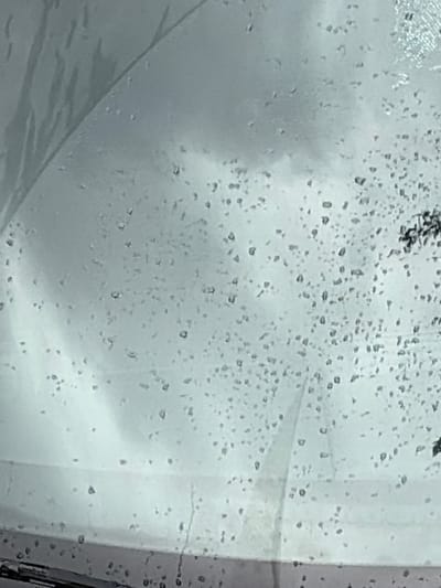 The image shows a close-up view of water droplets on a car windshield. The droplets are varied in size and distribution, with some appearing clustered and others spread out. The background is blurred and appears to be an overcast sky or a dimly lit environment. There is a dark, curved object at the bottom of the image, which is likely the windshield wiper. The overall impression is of a car driving in rainy or foggy conditions. The image does not provide enough visual information to determine its specific location in Grapevine, Texas, or to identify any people, actions, or interactions.