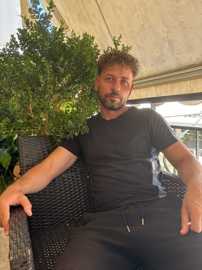 A man with curly brown hair and a beard sits in a dark wicker chair, partially obscured by a green leafy plant. He is wearing a black t-shirt with grey accents on the sleeves and black shorts. His left arm rests on the chair's armrest, while his right hand is on his lap. The background appears to be an outdoor seating area with a light-colored awning overhead. The man is looking directly at the camera. This scene is located in Ilion, Greece.