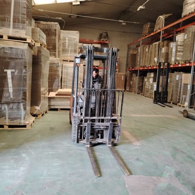 A man is driving a forklift in a warehouse with high ceilings. He is wearing a dark jacket and is looking forward. The forklift is orange and black, with a metal cage around the driver. The warehouse is filled with stacks of boxes, wrapped in plastic and stacked on pallets. The boxes are of various sizes and shapes, some are tall and narrow, while others are short and wide. The floor of the warehouse is concrete and appears to be worn, with scuff marks and some patches of repair. In the background, there are tall metal shelves filled with more boxes. The lighting in the warehouse is artificial, with fluorescent lights visible on the ceiling. The overall impression is one of an industrial setting, likely a storage facility for manufactured goods. The image does not provide enough visual information to determine the specific city or location within Рахманово, Russia.