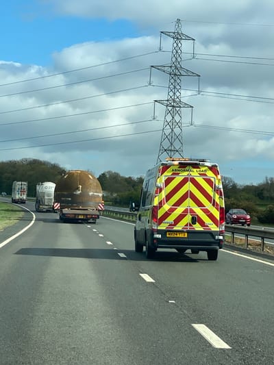 The image shows a convoy transporting a large, spherical, metal structure on a flatbed trailer along a highway. An "Abnormal Load" escort vehicle is positioned behind the trailer. The escort vehicle is a white van with red and yellow diagonal stripes on the rear and the words "ABNORMAL LOAD" displayed. The license plate of the van is visible and reads "NX24 YTB". The convoy is moving along a multi-lane highway marked with white lines. An electricity pylon is visible to the left of the convoy, and trees and greenery can be seen along the highway. The image was likely taken in Knayton, United Kingdom.