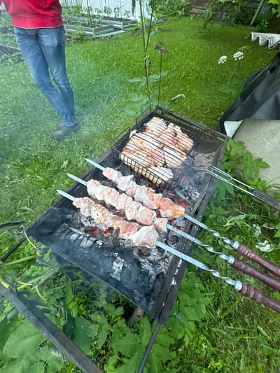 A top-down, eye-level shot captures a barbecue scene in a grassy yard in Stupino, Russia. The focus is on a black metal grill, filled with charcoal and glowing embers, supporting two cooking racks. On the lower rack, three metal skewers hold marinated meat, arranged in a row over the coals, with visible smoke rising. The upper rack contains a collection of raw chicken pieces, laid out in a grid pattern. To the left of the grill, a person's legs, clad in blue jeans and dark shoes, are visible, standing on the grass. The background features lush green grass, interspersed with plants and some white flowers. In the distance, a raised garden bed with plants is partially visible, suggesting a domestic outdoor setting. The overall ambiance is one of outdoor cooking and leisure.