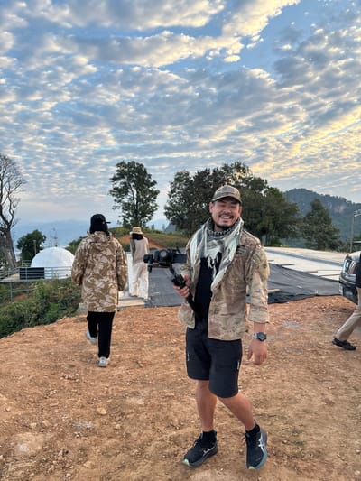 A man smiles, holding a camera stabilizer, dressed in camouflage shirt and cap, black shorts, and athletic shoes. Another person stands behind him, also in a camouflage jacket. In the background, a couple of individuals wearing white robes are visible. The setting appears to be outdoors, with a cloudy sky and mountainous terrain in the distance. There's a large white dome-shaped structure and some trees on the left side of the image. The ground is dry and dusty.