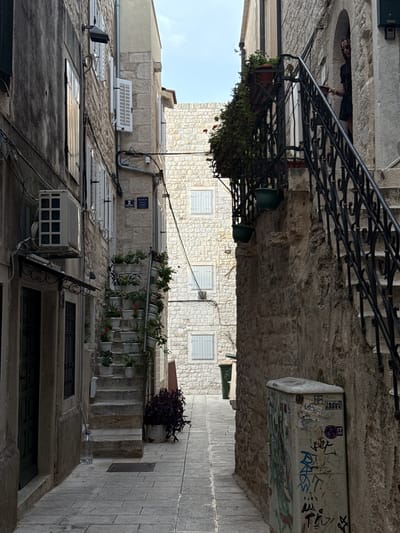The image shows a narrow, stone-paved alleyway in what appears to be an old European city. The buildings on either side are constructed from light-colored stone, with visible architectural details like windows with white shutters, balconies with wrought-iron railings, and air conditioning units. Greenery cascades from planters on several balconies, adding a touch of life to the scene. The alley slopes downwards and to the right, with a paved walkway on the right side of the frame.
In the upper left portion of the image, a person wearing dark clothing and sunglasses is partially visible, leaning over a balcony railing. The person appears to be observing the alley below. On a vertical surface on the right side of the alley, there is a rectangular object covered in graffiti.
The scene suggests a warm, sunny day, as indicated by the bright light and shadows. The architecture and the presence of narrow alleys are characteristic of historical cities, and the provided context specifies Split, Croatia. The overall mood is one of quiet observation and the passage of time in an ancient setting. There is some graffiti visible on a utility box in the upper right, but no other significant text is legible.