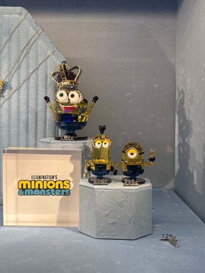 The image shows a retail display featuring crystal figurines of Minions characters from the "Illumination's Minions & Monsters" franchise. There are three figurines of different Minions characters on display, each with a price tag of 250.00. A fourth character is wearing a crown. The display also includes necklaces. The location is Spreitenbach, Switzerland.