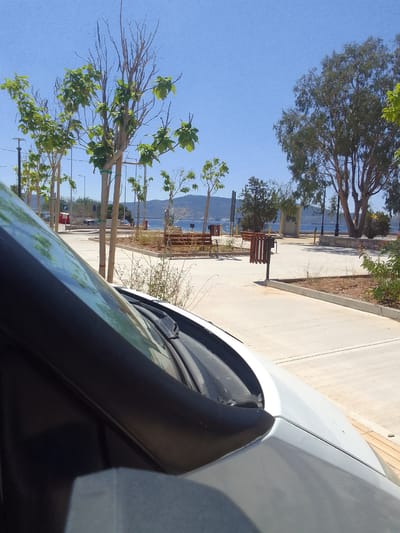 The image captures a scenic view of a waterfront area, likely within the Municipal Unit of Elefsina, Greece, on a clear, sunny day. In the foreground, the hood and windshield of a white vehicle are visible, partially obscuring the view. Beyond the vehicle, a paved walkway stretches towards the water, lined with young trees with sparse foliage and some taller, mature trees.

Several park benches and trash receptacles are strategically placed along the walkway, suggesting a public recreational space. In the background, the blue expanse of the sea meets a distant mountainous horizon. The presence of streetlights, a parking sign, and a red structure that could be a small kiosk or booth indicate urban development around the waterfront. The overall scene is tranquil and inviting, suggesting a pleasant day for outdoor activities.