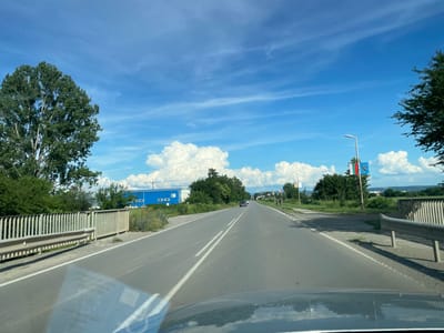 The image shows a road in Musachevo, Bulgaria, on a sunny day with a blue sky and white clouds. The perspective is from inside a vehicle, looking forward. On the left side of the road, there is a metal fence and a large tree. Further down the road on the left, there is a long, blue building with white-framed windows. On the right side of the road, there are more trees, a lamppost with flags, and a few people walking. The road has two lanes in each direction, marked by white lines. There are a few cars on the road. The overall environment appears to be a semi-urban or suburban area.