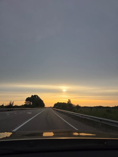 The image shows a highway at sunset. The road stretches into the distance with a white line dividing the lanes. There is a guardrail on the right side of the road. The sky transitions from gray to orange, with the sun visible on the horizon. Trees are visible along the side of the road. A portion of a vehicle is visible in the lower right corner of the image. It is not possible to confirm the location is in Aur, Russia based on visual information alone.