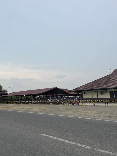 The image depicts an outdoor parking area, likely in Glokaron, Indonesia, featuring a row of covered parking stalls filled with motorcycles. The stalls are covered by a long, maroon-colored roof supported by dark posts, and are enclosed by a chain-link fence with yellow and black striped posts. Beyond the stalls, a building with a similar maroon roof and beige walls is visible, equipped with an air conditioning unit. In the foreground, a paved road runs horizontally across the frame, with a white line marking the edge. The sky is overcast with a pale blue hue. No people are clearly visible in the image, but their presence is suggested by the numerous parked motorcycles.