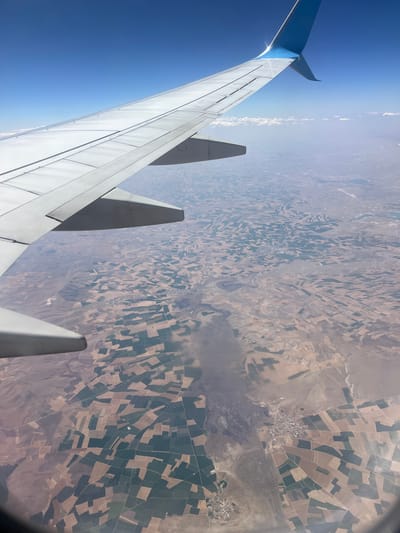 An aerial view from an airplane window shows a vast, arid landscape dotted with numerous rectangular agricultural fields. The fields exhibit a patchwork of brown, tan, and green, indicating different crops or stages of cultivation. A small town, likely in the vicinity of Eğirdir, Turkey, is visible in the center of the image, with clusters of buildings and roads. The airplane's wing, predominantly white with a blue tip, occupies the upper left portion of the frame, casting a shadow over some of the terrain. The sky above is a clear, bright blue.