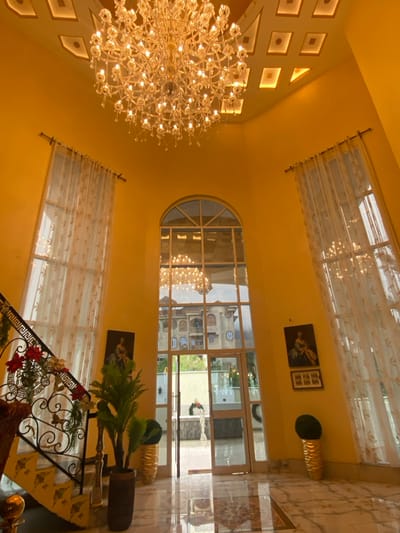 The image shows the interior of a grand, modern house in Buea, Cameroon. The walls are painted a bright yellow and adorned with decorative moldings. A large, ornate chandelier hangs from the center of the ceiling. There are two large windows flanking a large glass door which leads to an outdoor area. White lace curtains hang from the windows. Two potted plants and a small table are placed at the base of each window. A black wrought iron banister and stairway are located on the left side of the room. An artwork depicting a female figure in a dress is hung on the wall. Two framed pictures of landscapes are hung above it. The floor is made of white marble tiles. The outside view shows a building with a multi-pane glass window and a fountain in a garden. The overall scene is one of luxury and sophistication.