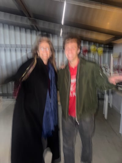 The image depicts two individuals, an adult female-presenting person and a young adult male-presenting person, posing side-by-side in an indoor setting. The entire scene is significantly motion-blurred. The specified location is Décines-Charpieu, France, though this detail is not visually verifiable within the image.
The female-presenting individual on the left wears a dark, possibly black, long coat or cape, a blue scarf draped around the neck, and eyeglasses. Long, light-colored hair is visible, and a gold-toned accessory appears near her left shoulder. She is smiling and looking towards the camera. Her right arm is obscured but appears to be positioned behind the male-presenting individual.
The male-presenting individual on the right wears a dark green bomber jacket over a red t-shirt featuring indistinct white graphics, and dark pants. He is also smiling and looking towards the camera. His left arm is extended outwards, away from his body, with the palm open.
The background features light-colored, corrugated metal paneling. Overhead artificial lighting is visible, creating prominent vertical light streaks due to the motion blur. On the far right, indistinct objects, possibly stacked chairs and other items including yellow shapes, are present on a light-colored surface. The floor appears to be a uniform, light-toned surface.