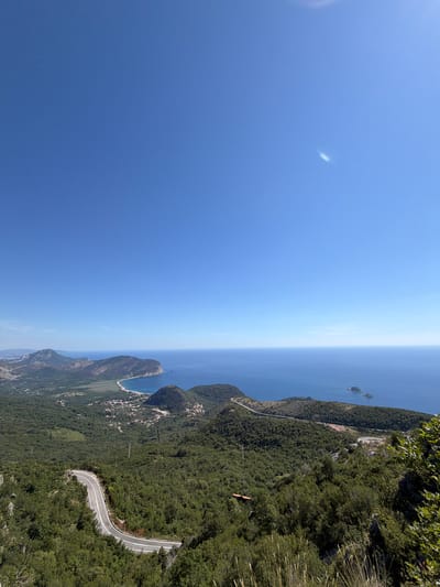 The image depicts a panoramic view of a coastal mountainous region under a clear blue sky with a wispy cloud formation. The foreground is dominated by a winding road cutting through dense green foliage. The road features white lane markings and appears to be a paved asphalt surface. In the mid-ground, a valley opens up, revealing a small settlement with red-tiled roofs nestled amongst the green landscape. Further in the distance, the coastline meets the deep blue sea, with a curved beach visible. Several peninsulas and hilly terrains extend into the sea. Two small islands are situated offshore, with a boat visible in the water near them. The overall scene suggests a natural landscape with a human presence in the form of a road and a small village. The location is identified as Novoselje, Montenegro.