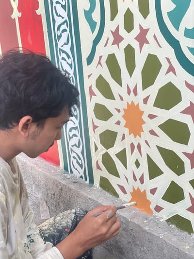 A person is painting an intricate geometric pattern on a ceiling. The design features interlocking shapes and stars in colors like green, red, turquoise, orange, and beige. The artist is focused on filling in an orange star with a paintbrush. A paint bucket rests nearby. The elaborate pattern suggests a cultural or architectural significance.